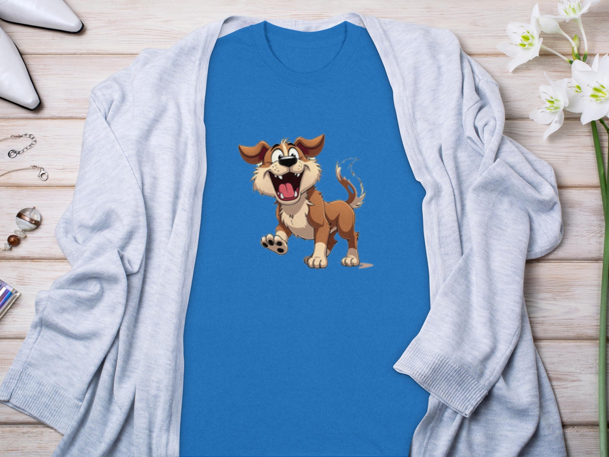 A Garment Graphics blue cartoon dog T-shirt, ideal for dog lovers, is displayed on a wooden surface, partially covered by a light gray cardigan. Nearby are white high heels, a silver bracelet, and white flowers, creating a charming ensemble for any occasion.