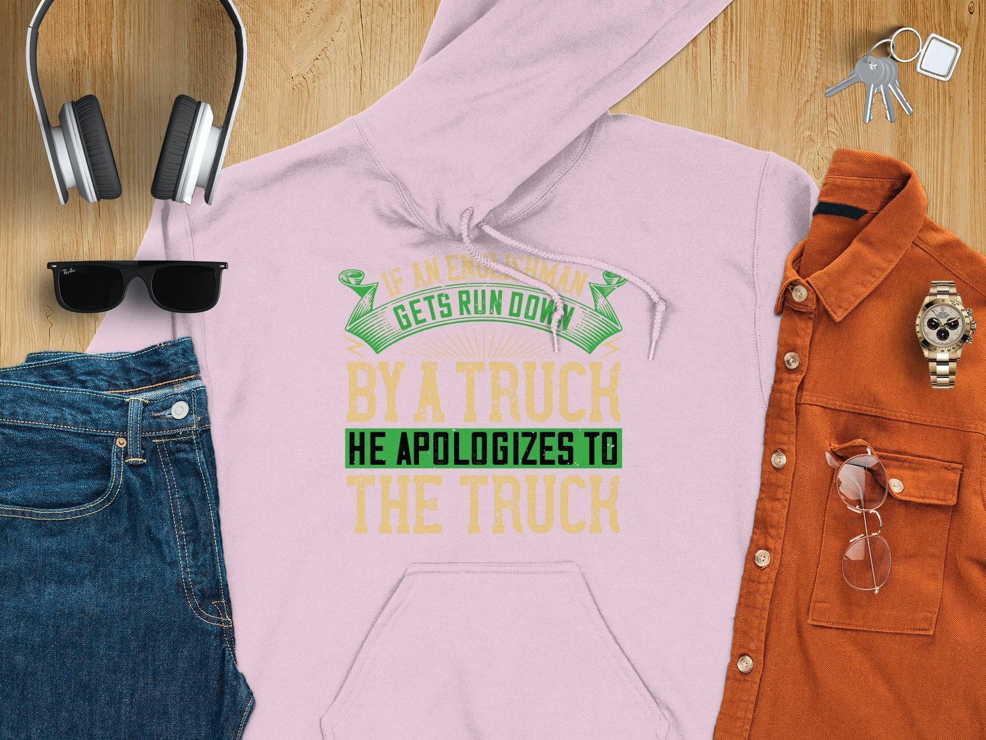 A flat lay includes a light pink humorous hoodie by Garment Graphics with the text, If an Englishman gets run down by a truck he apologizes to the truck, reflecting English etiquette, surrounded by headphones, sunglasses, jeans, an orange shirt, and a wristwatch on a wooden surface.