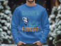 A person wearing Garment Graphics blue graphic sweatshirt features a fish, someone fishing in a boat, and Daddys Fishing Buddy in vibrant colors. Perfect for outdoor lovers, with snowy trees forming the blurred background.