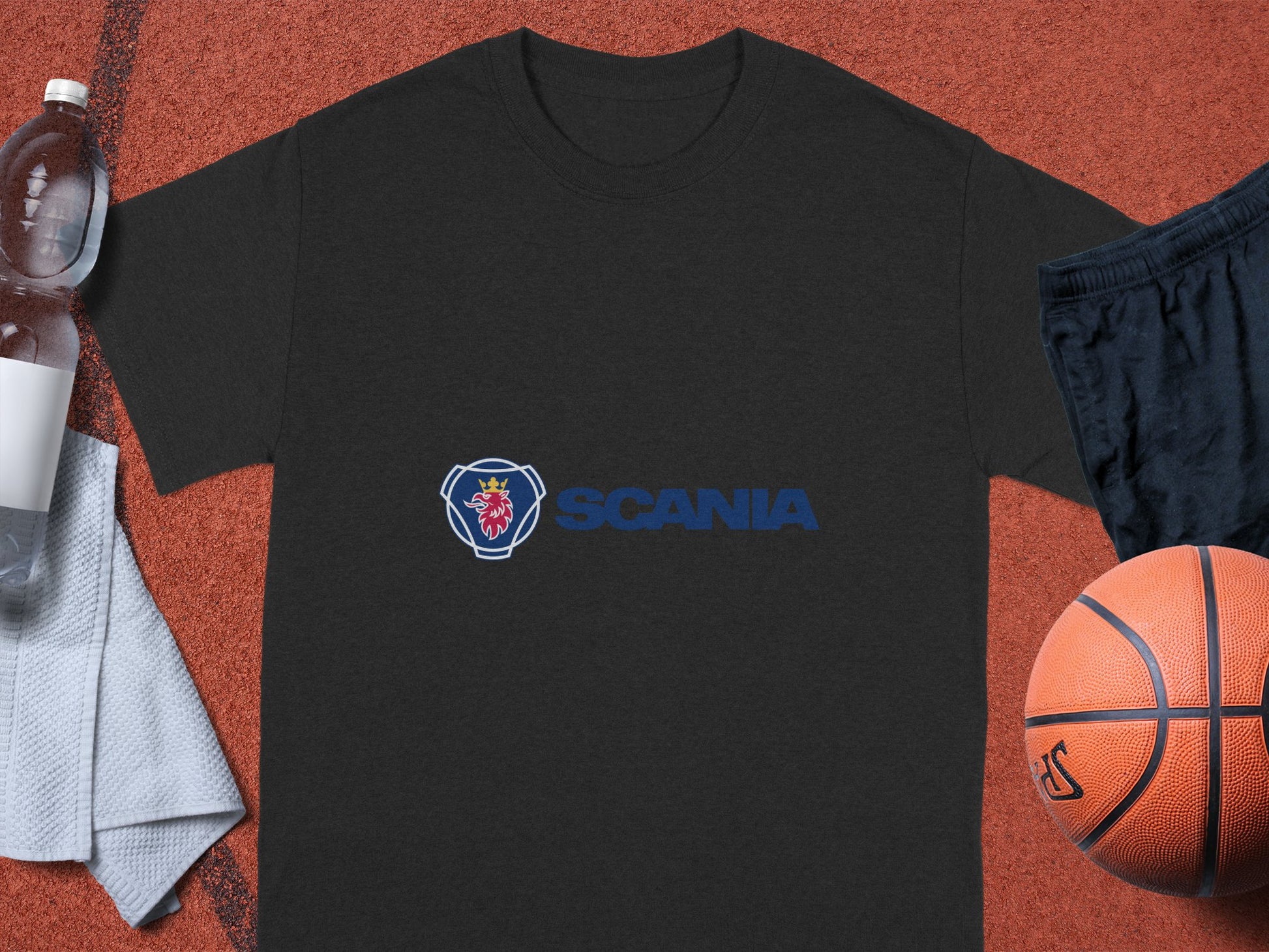 A black cotton T-shirt with the Scania logo by Garment Graphics rests on a sports court. A basketball is on the right with black shorts, while a partially seen white towel on the left holds a half-full water bottle—ideal for Scania fans.
