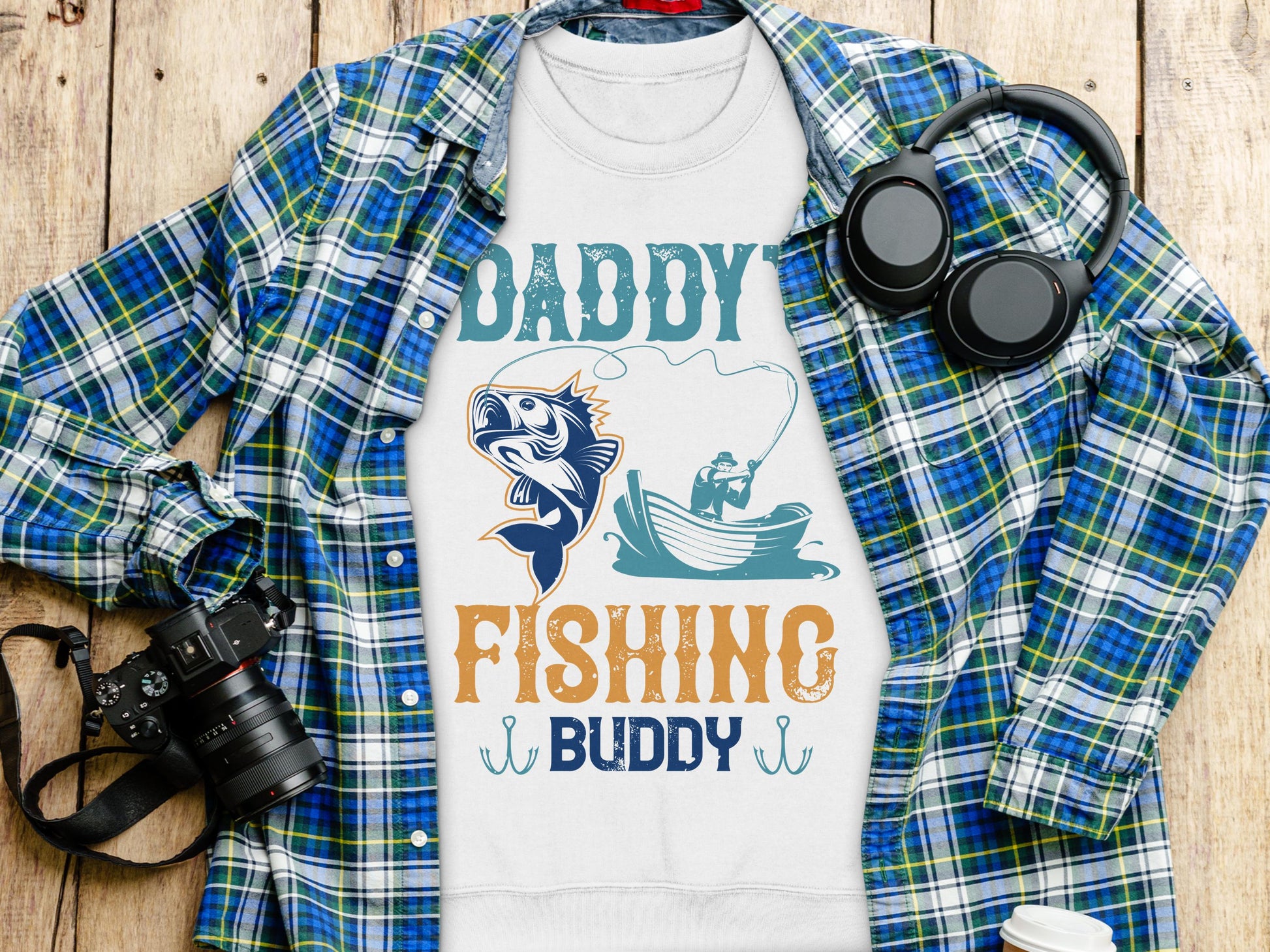 The Garment Graphics white graphic sweatshirt with Daddys Fishing Buddy text features a fisherman and fish illustration. Its displayed on a blue-green plaid shirt alongside a camera and black headphones, making it ideal for outdoor enthusiasts.