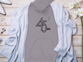 A Garment Graphics gray 45 RPM hoodie for vinyl enthusiasts lies on a wooden surface, with a light gray cardigan draped over it. Nearby are white high heels, jewelry pieces like bracelets and rings, and white flowers.