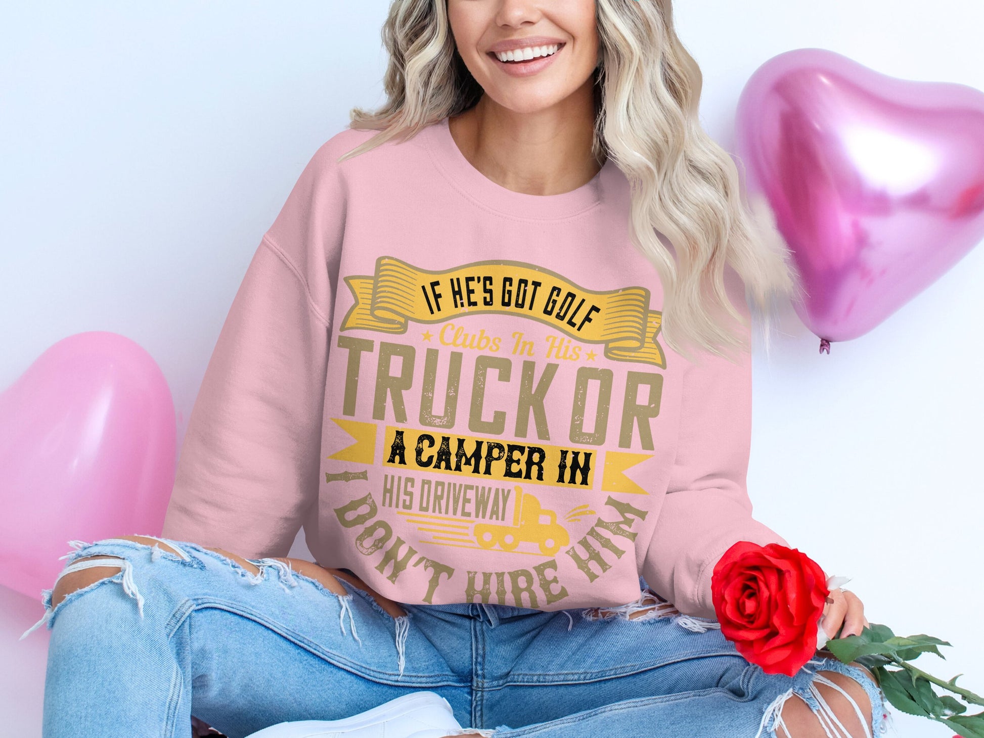 A person with wavy blonde hair sits on the floor wearing a Garment Graphics casual golf clubs sweatshirt with a playful message about golf, a truck, and a camper. Holding a red rose, they are surrounded by pink heart-shaped balloons.
