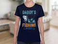 A person wears a navy blue Garment Graphics fishing t-shirt with the text Daddys Fishing Buddy and illustrations of a fish and fishing boat. The softly blurred indoor setting accentuates the playful charm of this shirt.