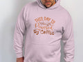 A stylish coffee enthusiast wears a light pink Garment Graphics hoodie featuring This Day is Powered by Coffee in brown and orange fonts. With their head slightly tilted down and hands in the pocket, its the perfect look for devoted fans of Coffee Hoodies.