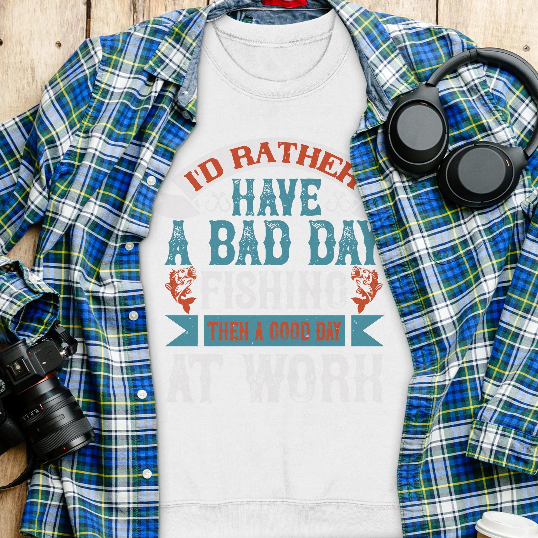 A white Garment Graphics t-shirt with Id rather have a bad day fishing than a good day at work sits on a plaid shirt. Nearby, headphones and a camera lie on the wooden surface—ideal for fishing fans who prefer unisex sizing in their casual wear.