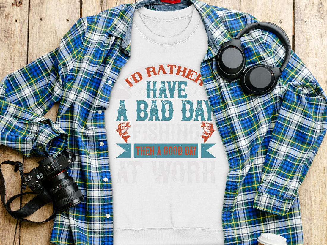 A white Garment Graphics t-shirt with Id rather have a bad day fishing than a good day at work sits on a plaid shirt. Nearby, headphones and a camera lie on the wooden surface—ideal for fishing fans who prefer unisex sizing in their casual wear.