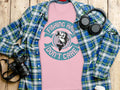 A distinctive Garment Graphics tee with a fish and the slogan Fishing Hair Dont Care is placed on a blue and green plaid shirt. Nearby are headphones, a camera, and a white cup on the wooden surface, ideal for everyday adventures.