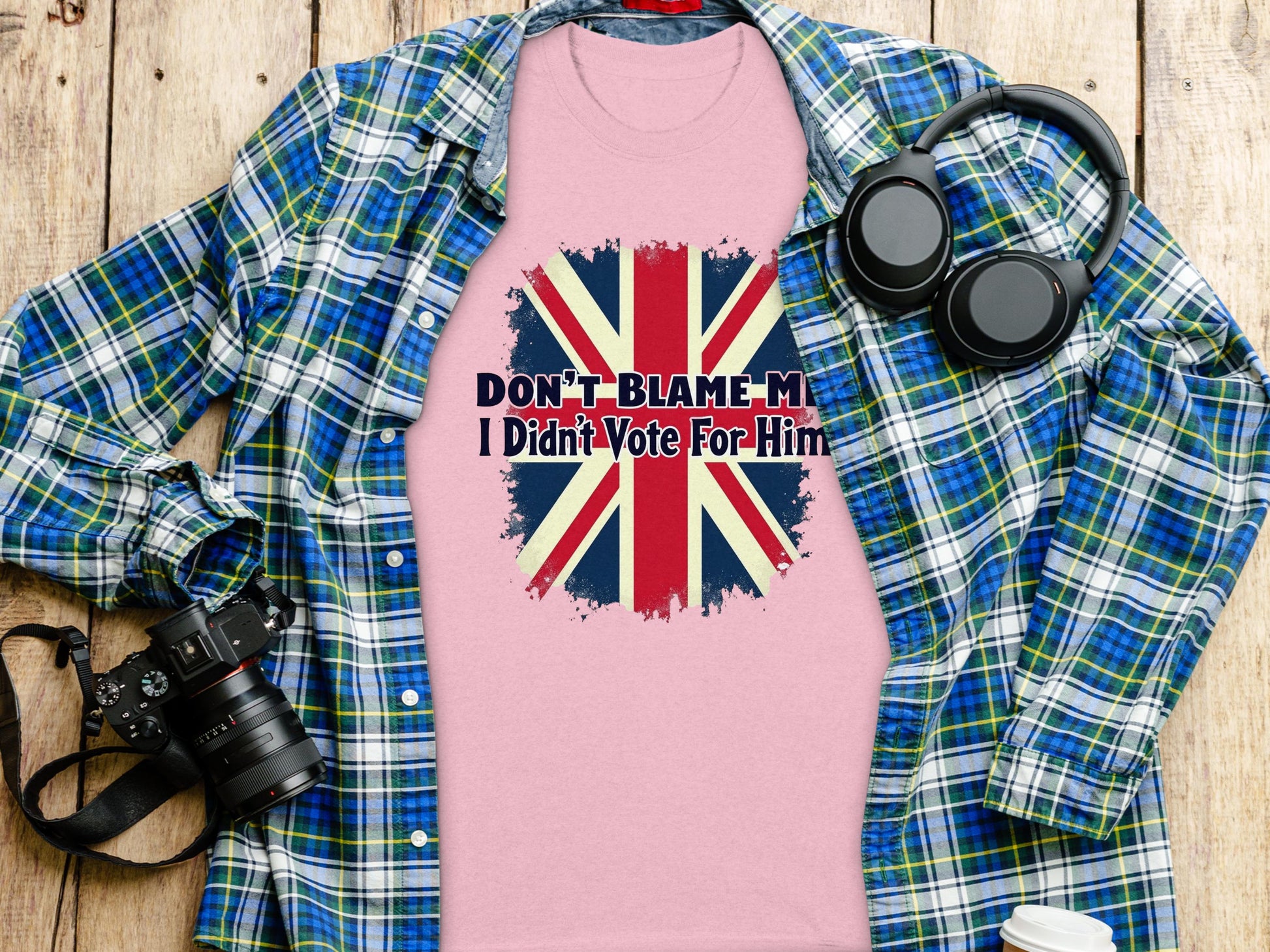 A bold statement pink T-shirt by Garment Graphics features a Union Jack and the text Dont Blame Me I Didnt Vote For Him, displayed on a wooden surface with a blue plaid shirt, camera, headphones, and white mug.