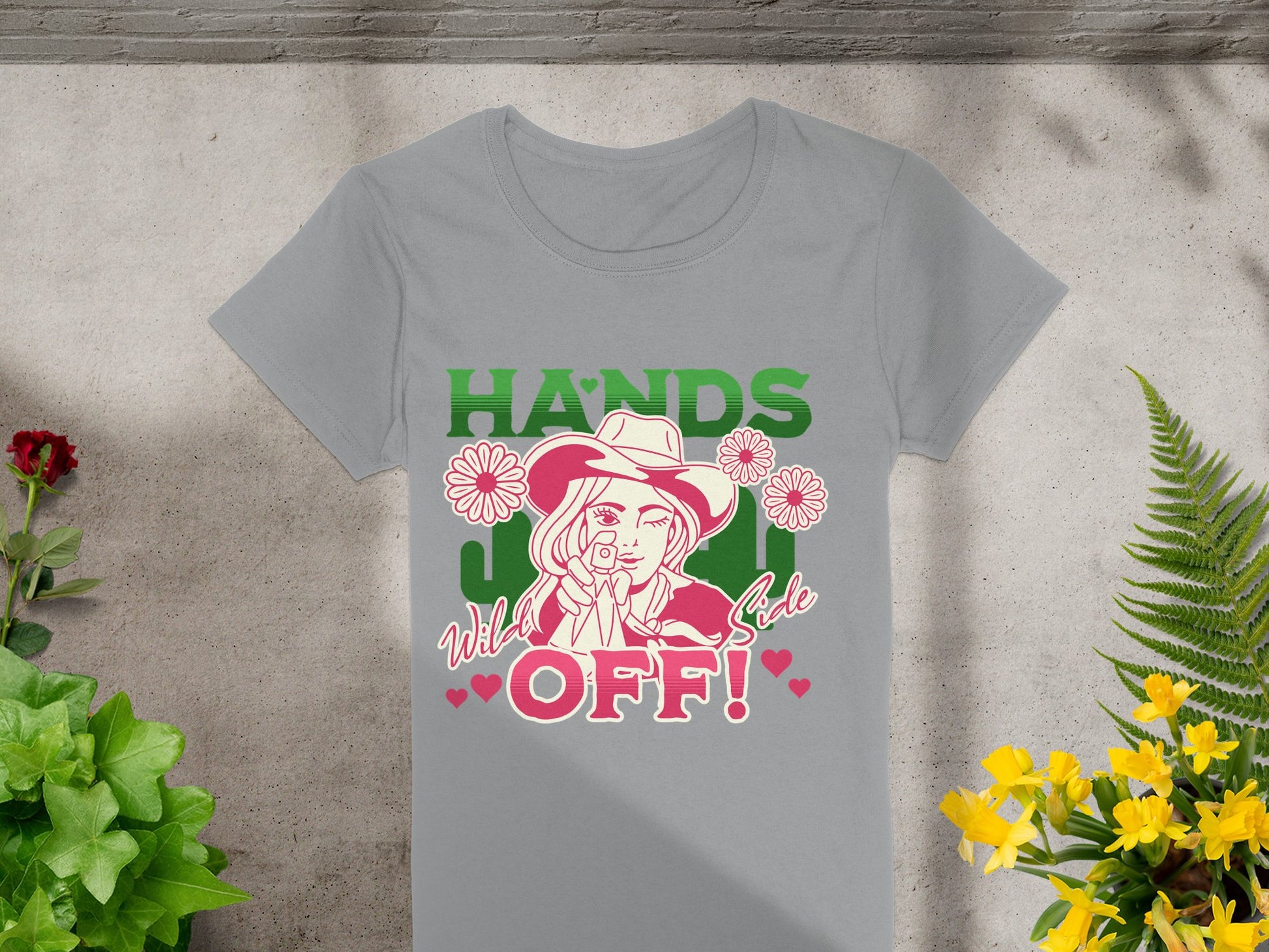 A unique Garment Graphics gray T-shirt boldly showcases a person in a cowboy hat surrounded by flowers and hearts. HANDS and OFF! burst above in green and pink, with Wild side also featured. Displayed on concrete next to plants, this stylish tee exudes flair.