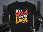 The Garment Graphics unisex Graphic Art Sweatshirt, featuring the vibrant message Its a Good Day to Laugh, is set on a gray background and paired with a smartphone, smartwatch, sneakers, dumbbells, and portable speaker to complement an active lifestyle.
