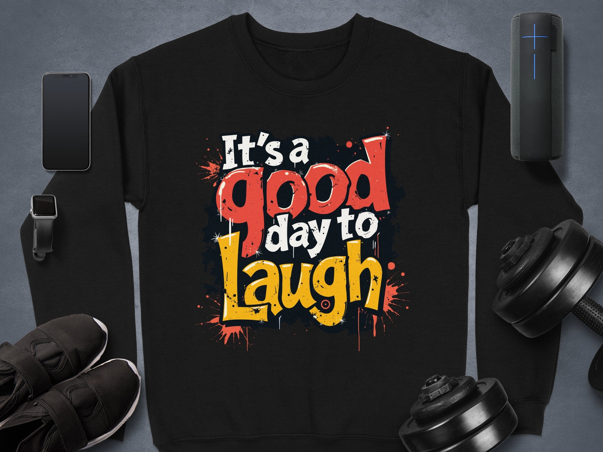 The Garment Graphics unisex Graphic Art Sweatshirt, featuring the vibrant message Its a Good Day to Laugh, is set on a gray background and paired with a smartphone, smartwatch, sneakers, dumbbells, and portable speaker to complement an active lifestyle.