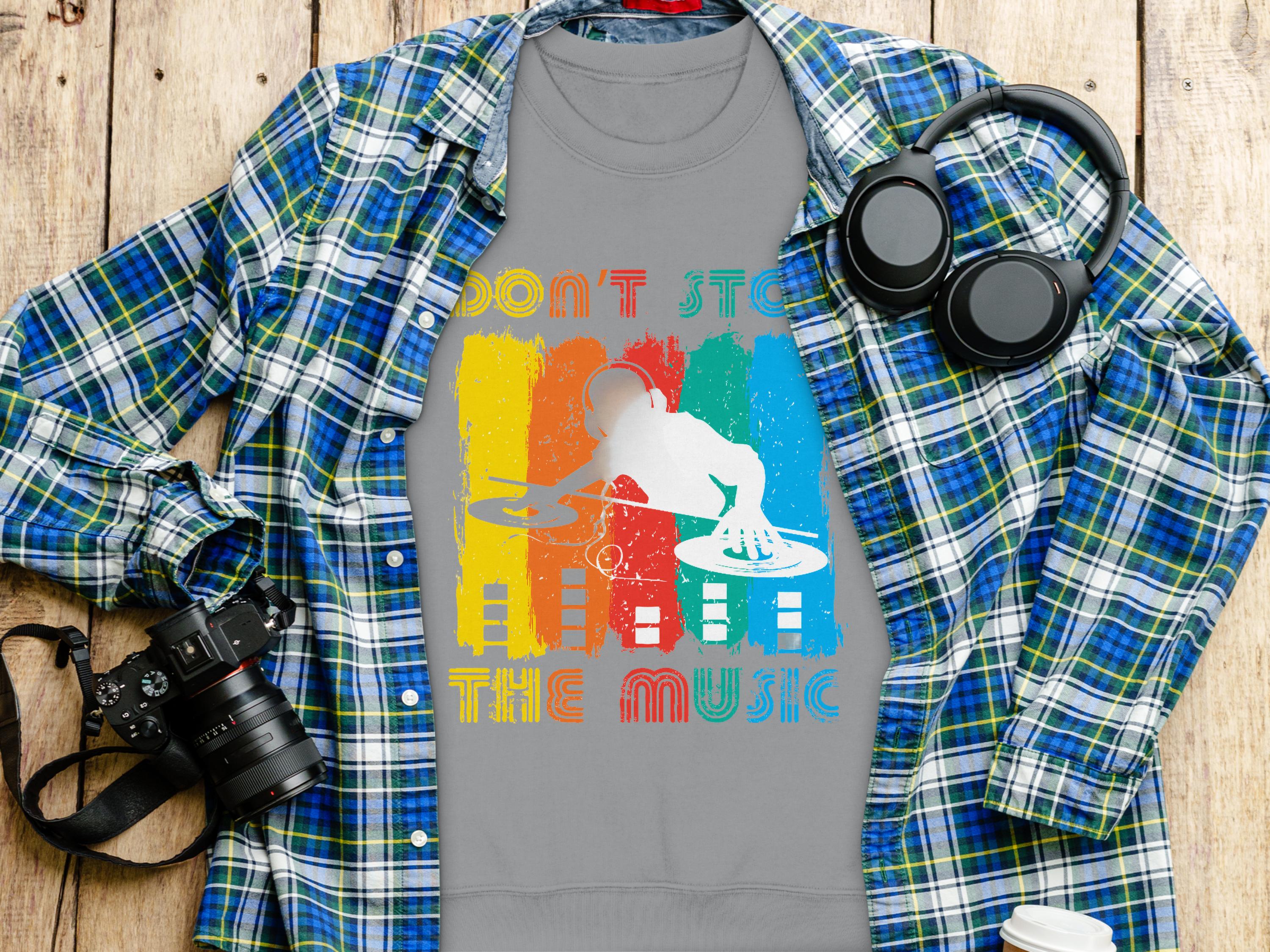 A gray Garment Graphics DJ T-shirt with Dont Stop the Music text peeks from under a blue and green plaid shirt. Ideal for music lovers, the setup includes headphones, a camera, and a coffee cup on wood. Perfect for unisex sizing fans who dont want to miss the beat.