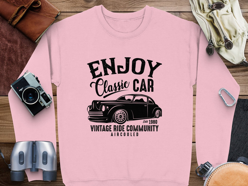 A Garment Graphics pink sweatshirt displays a vintage car graphic with the text Enjoy Classic Car, Vintage Ride Community Aircooled, and Est 1980. Surrounded by retro items, its ideal for classic car enthusiasts and lovers of timeless fashion.