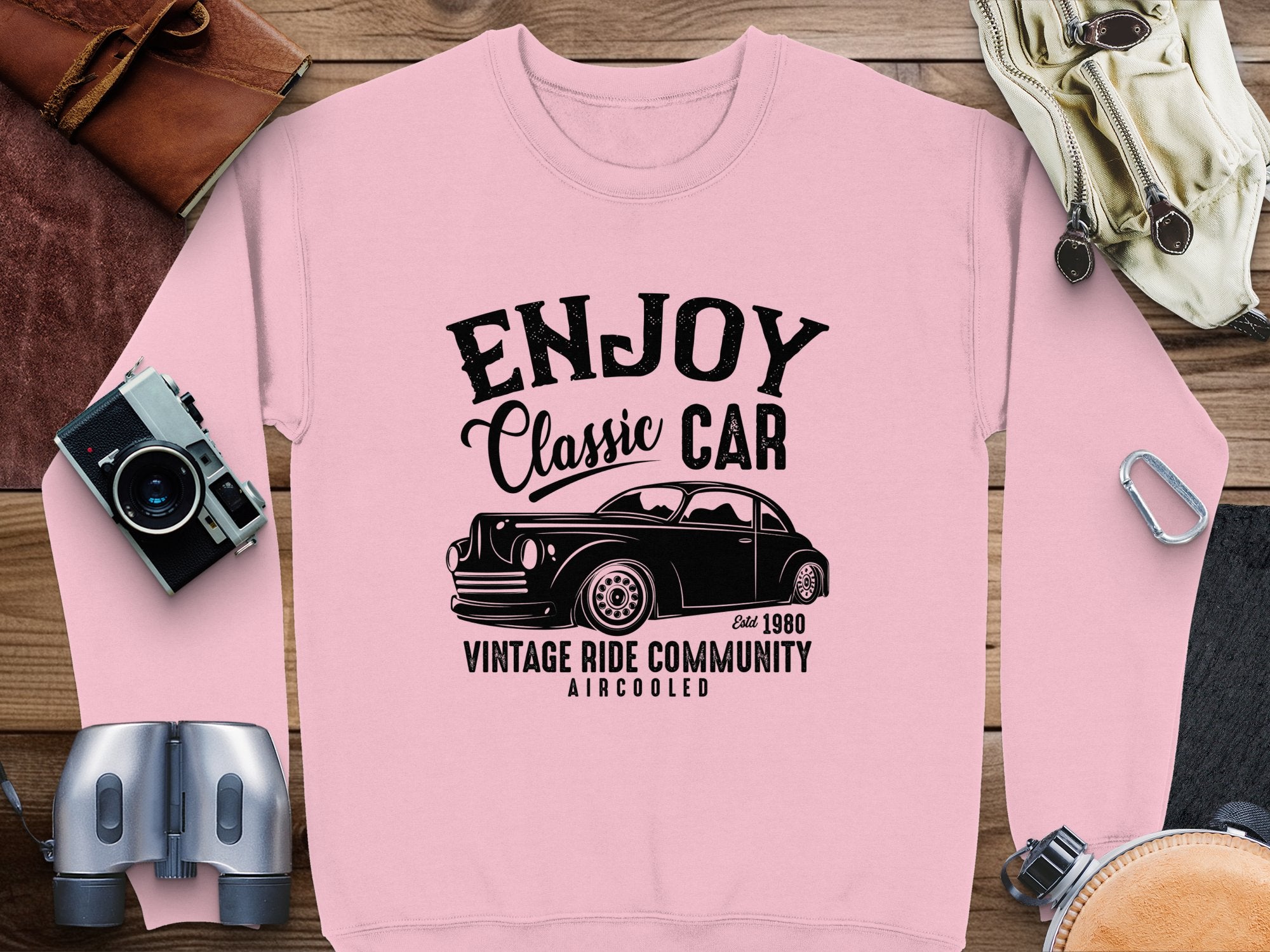 A Garment Graphics pink sweatshirt displays a vintage car graphic with the text Enjoy Classic Car, Vintage Ride Community Aircooled, and Est 1980. Surrounded by retro items, its ideal for classic car enthusiasts and lovers of timeless fashion.