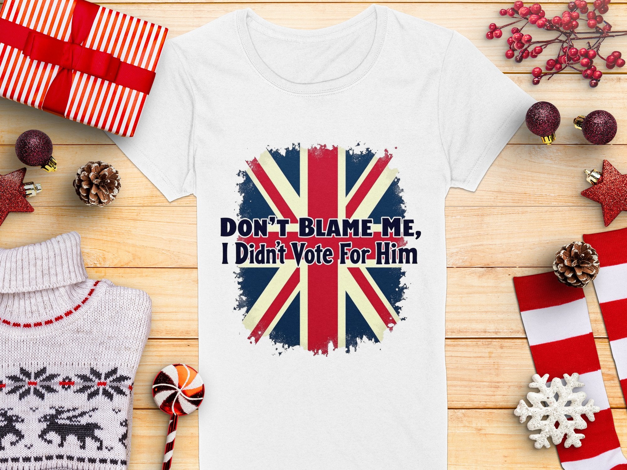 The Garment Graphics white t-shirt, made from 100% cotton, features the Union Jack and Dont Blame Me, I Didnt Vote For Him. Amid wrapped gifts, red and white decorations, a knitted sweater, and a candy cane on a wooden surface,this shirt stands out boldly.