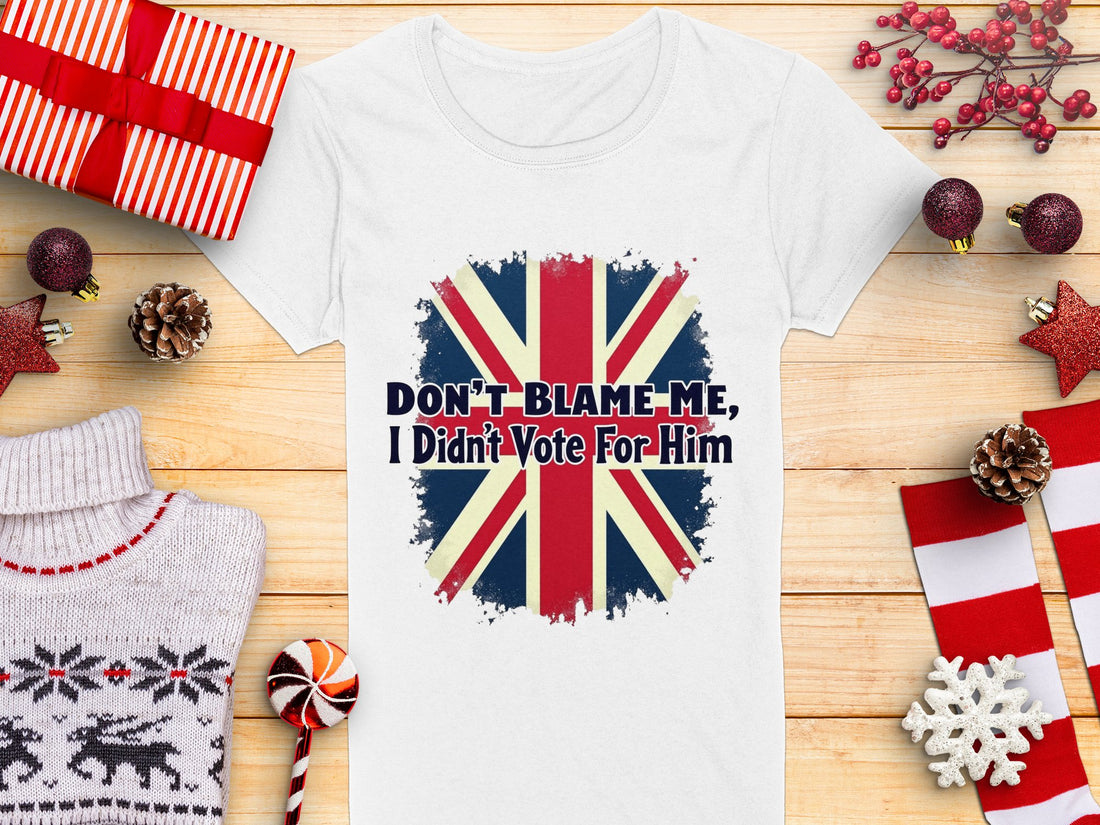 The Garment Graphics white t-shirt, made from 100% cotton, features the Union Jack and Dont Blame Me, I Didnt Vote For Him. Amid wrapped gifts, red and white decorations, a knitted sweater, and a candy cane on a wooden surface,this shirt stands out boldly.