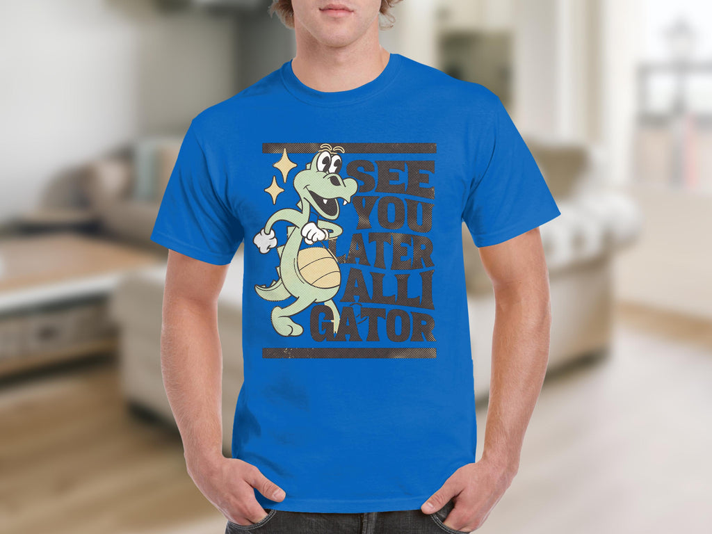 Sporting casual wear, a person wears a Garment Graphics blue T-shirt featuring a cartoon alligator with a basketball, captioned SEE YOU LATER ALLIGATOR, set against blurred interior and neutral-toned furniture.