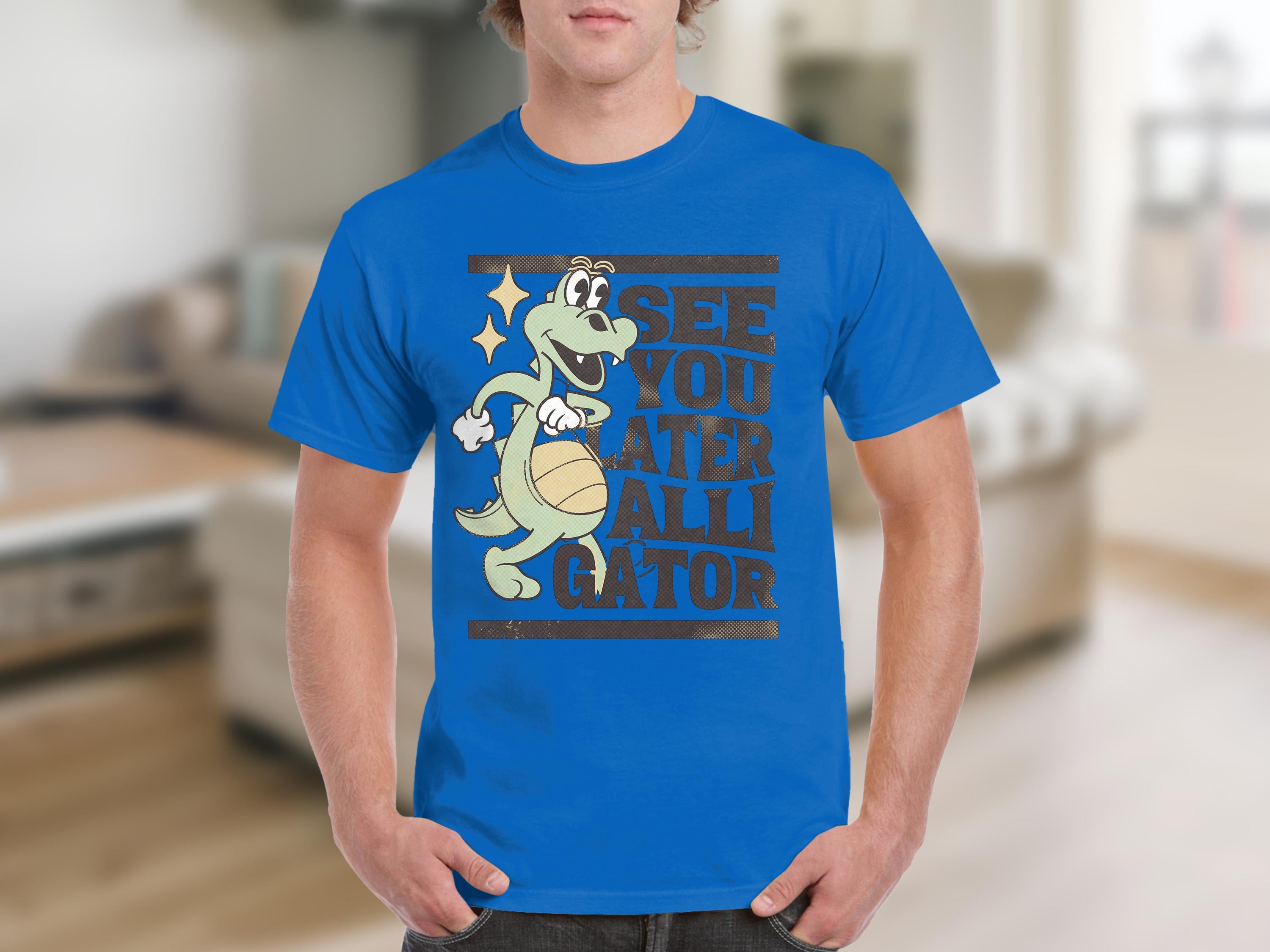 Sporting casual wear, a person wears a Garment Graphics blue T-shirt featuring a cartoon alligator with a basketball, captioned SEE YOU LATER ALLIGATOR, set against blurred interior and neutral-toned furniture.