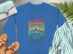 Garment Graphics blue sweatshirt displays a vibrant Woodstock 1969 bird and flower design, exuding retro vibes. Its styled with tan sandals, a striped shirt, wristwatch, and mint purse on a gray surface adorned with green ferns.