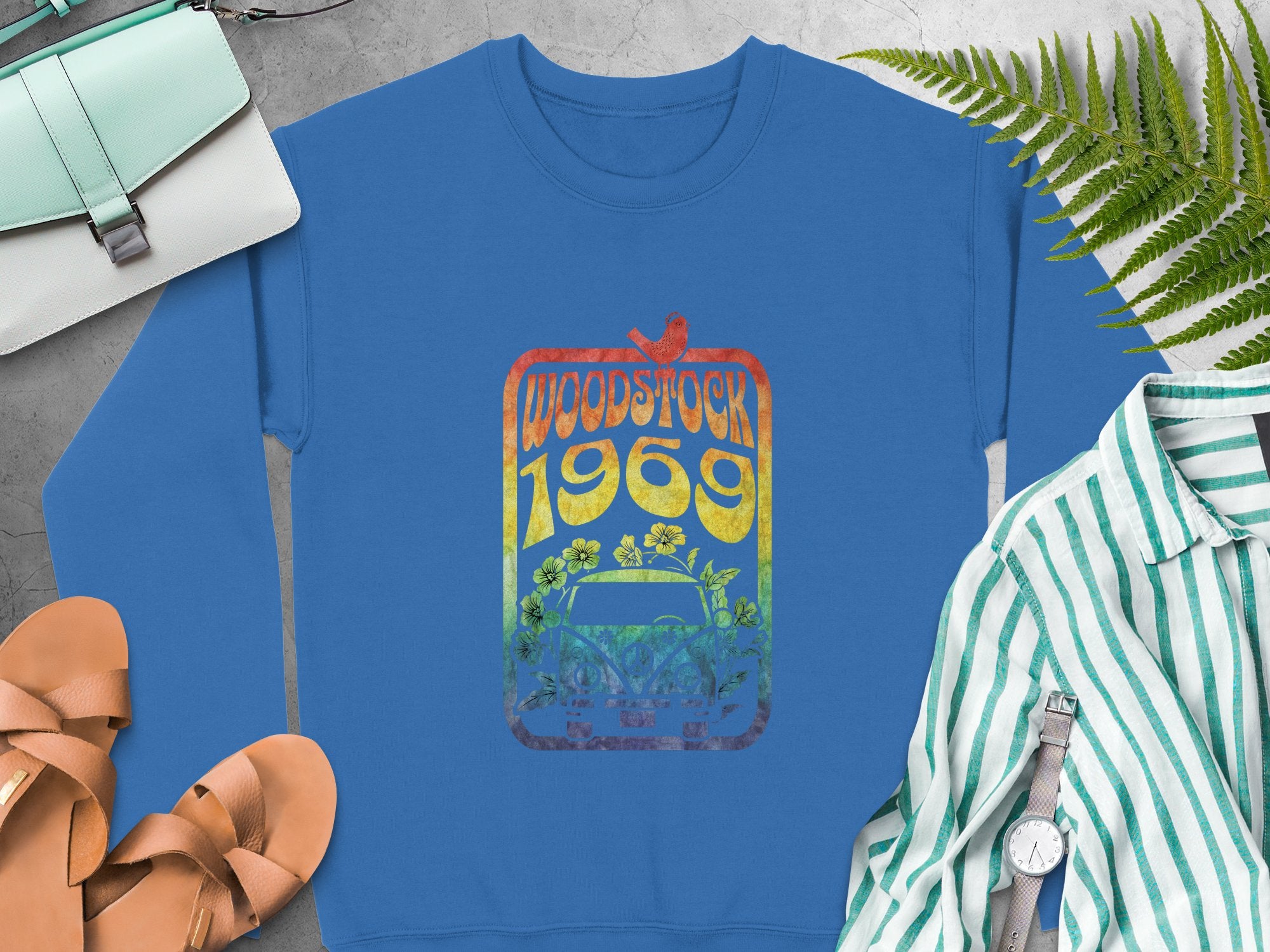 Garment Graphics blue sweatshirt displays a vibrant Woodstock 1969 bird and flower design, exuding retro vibes. Its styled with tan sandals, a striped shirt, wristwatch, and mint purse on a gray surface adorned with green ferns.