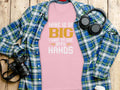 A Garment Graphics pink graphic tee with the design Mine is so big I have to use both hands sits on a wooden surface under a classic fit blue and green plaid shirt, surrounded by a camera, headphones, and a cup.