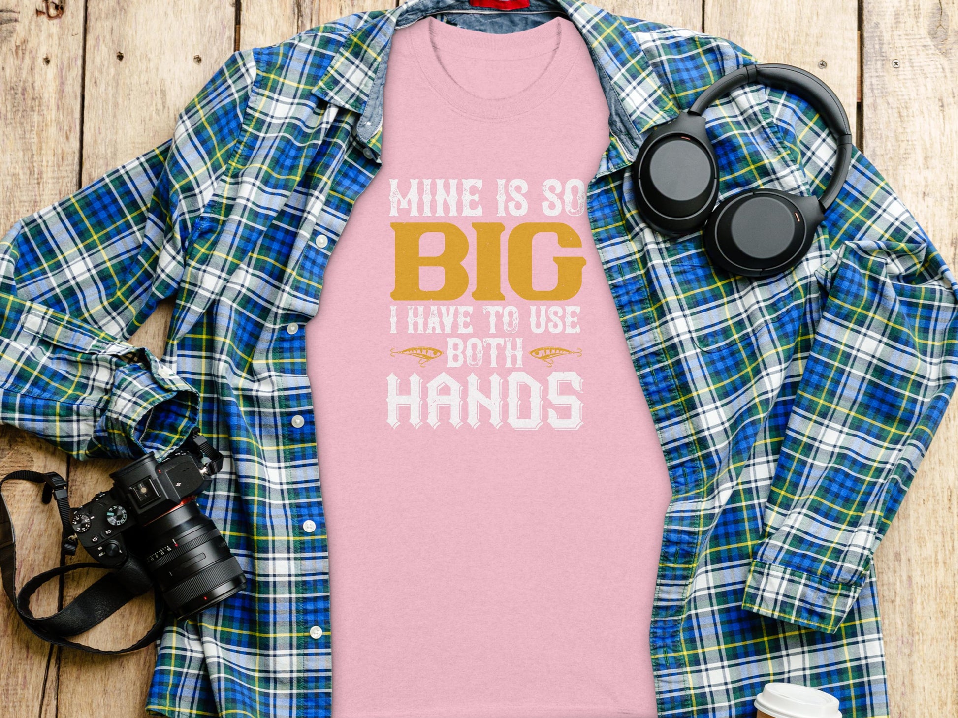 A Garment Graphics pink graphic tee with the design Mine is so big I have to use both hands sits on a wooden surface under a classic fit blue and green plaid shirt, surrounded by a camera, headphones, and a cup.