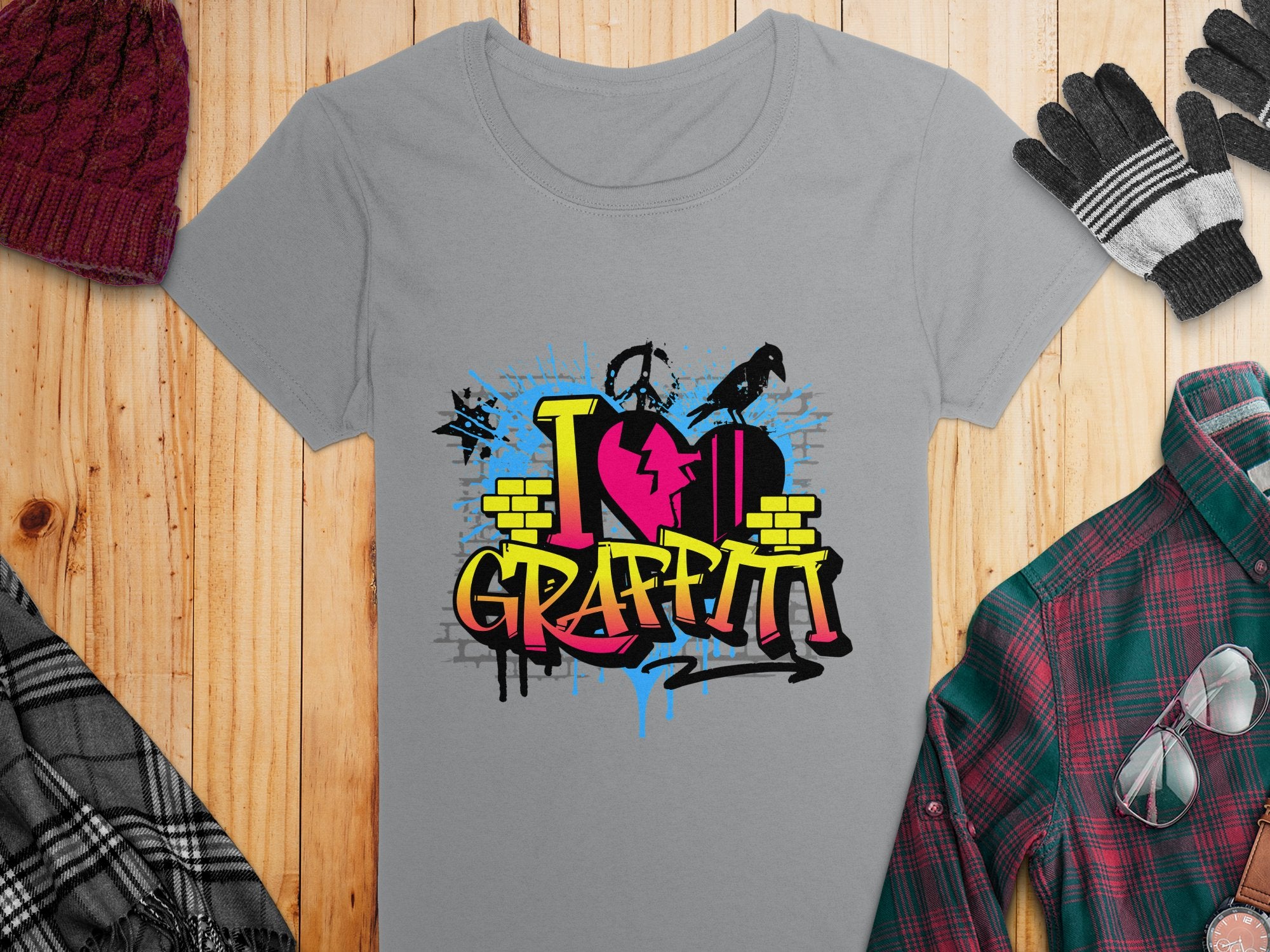 Garment Graphics gray t-shirt features a vibrant I Love Graffiti design with graffiti text and brick patterns, ideal for street art fans. Displayed on wood, it pairs well with a maroon knit hat, gray gloves, and a green plaid shirt.