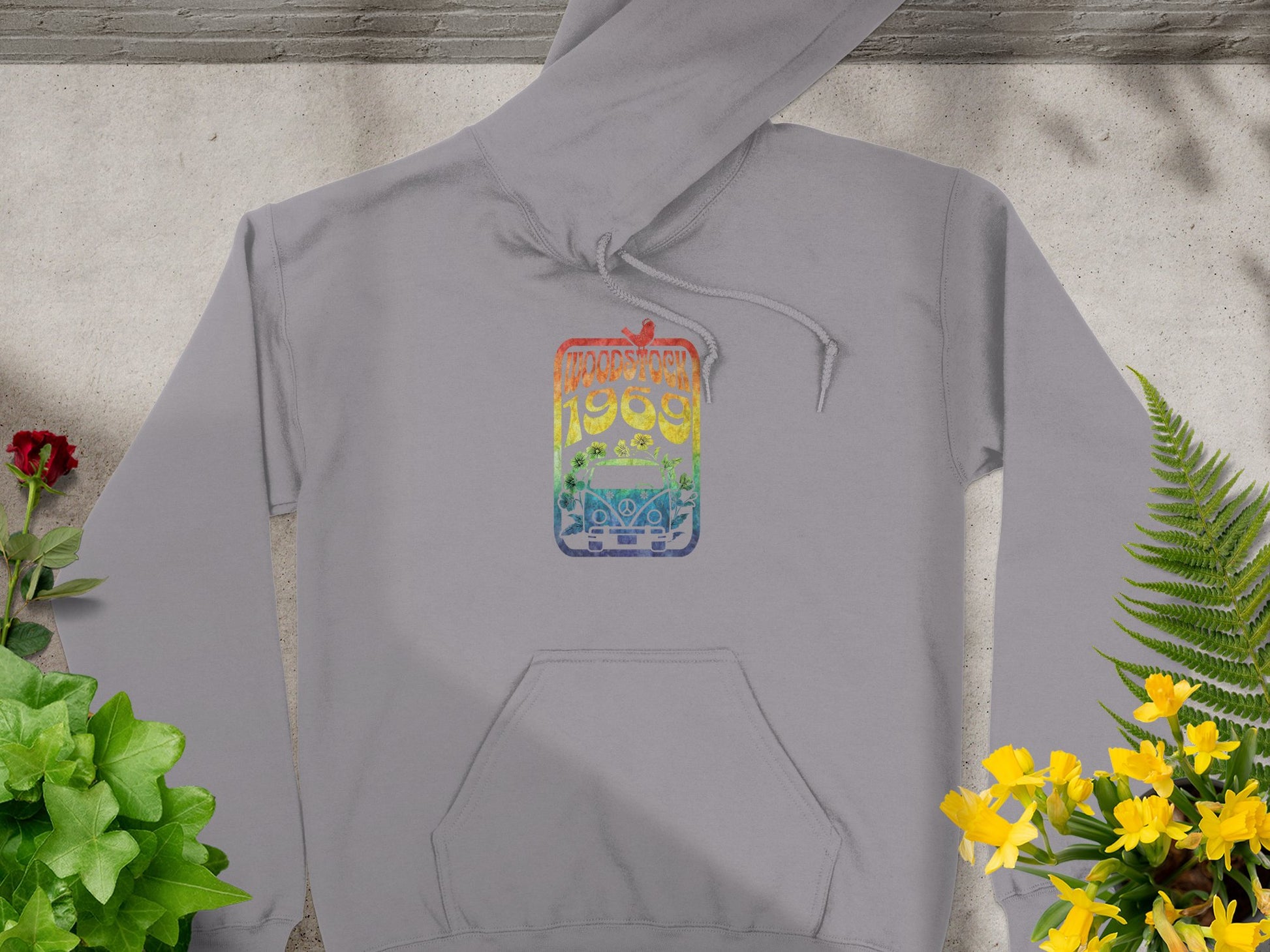 Garment Graphics gray hoodie features a vibrant 1980 graphic on the chest, photographed outdoors against a textured backdrop. Its retro Woodstock 1969 vibe is complemented by surrounding green, red, and yellow plants.