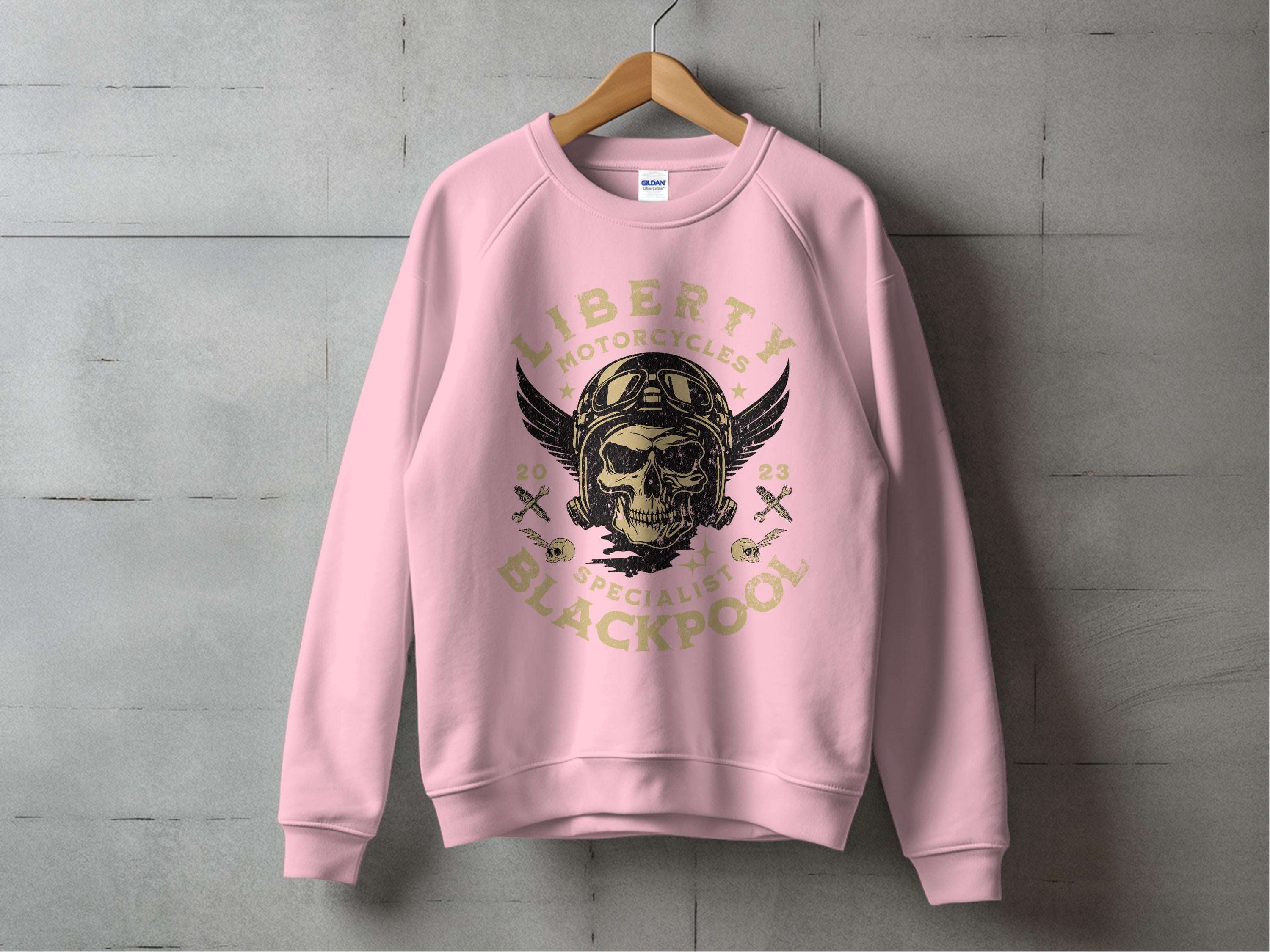 A pink sweatshirt hangs on a concrete wall, featuring a unique winged skull and helmet design. It reads Liberty Blackpool Motorcycle Specialist 2023, making it stand out from the Garment Graphics Blackpool 2023 Sweatshirts collection.