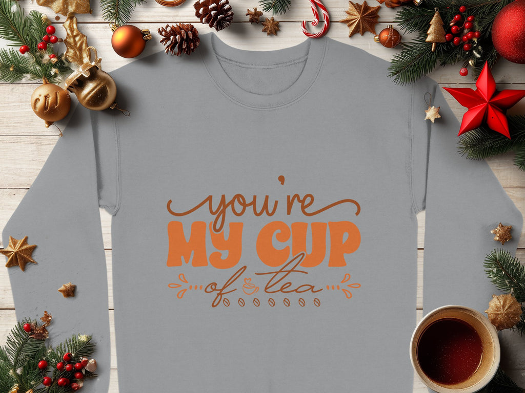 A cozy gray Garment Graphics sweatshirt with Youre My Cup of Tea in orange sits on a wooden surface. Perfect for tea lovers, it is adorned with festive Christmas decorations, pine branches, pinecones, ornaments, and a steaming cup of tea nearby.