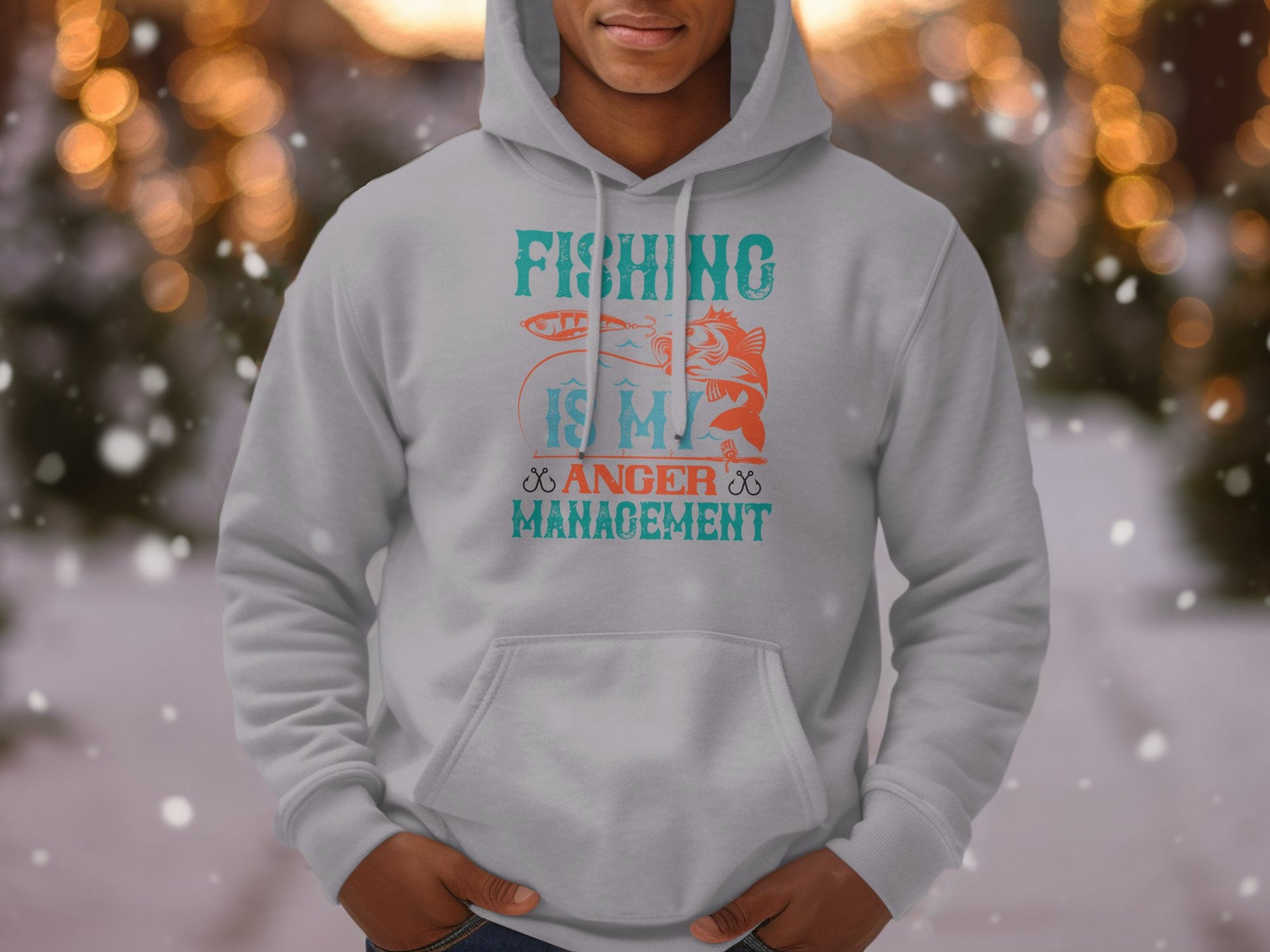 A person wears a Garment Graphics Anglers Wardrobe gray hoodie, displaying Fishing is My Anger Management in orange and teal with fish and rod graphics, set against a blurred lights and snowy backdrop.