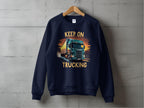 A navy blue graphic sweatshirt from Garment Graphics hangs against a concrete wall, featuring a bold truck design and the phrase Keep On Trucking in striking yellow letters.