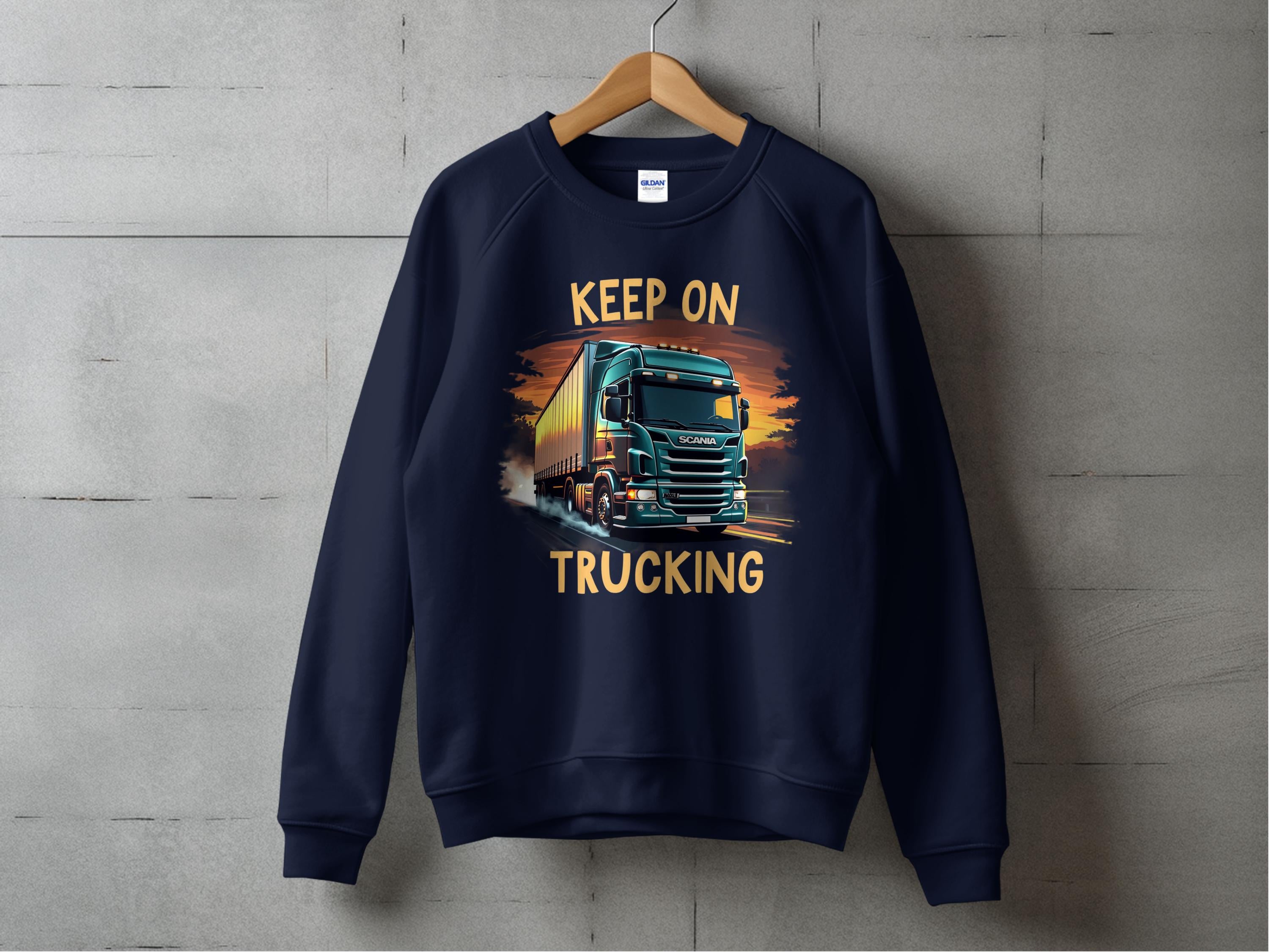A navy blue graphic sweatshirt from Garment Graphics hangs against a concrete wall, featuring a bold truck design and the phrase Keep On Trucking in striking yellow letters.