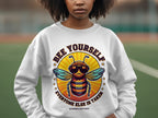 A person wears a Garment Graphics white summer edition sweatshirt with a bee in sunglasses. The top text says Bee Yourself, and below, it reads, Everyone Else Is Taken. Available in unisex sizes, the fun design stands out against a blurred background.