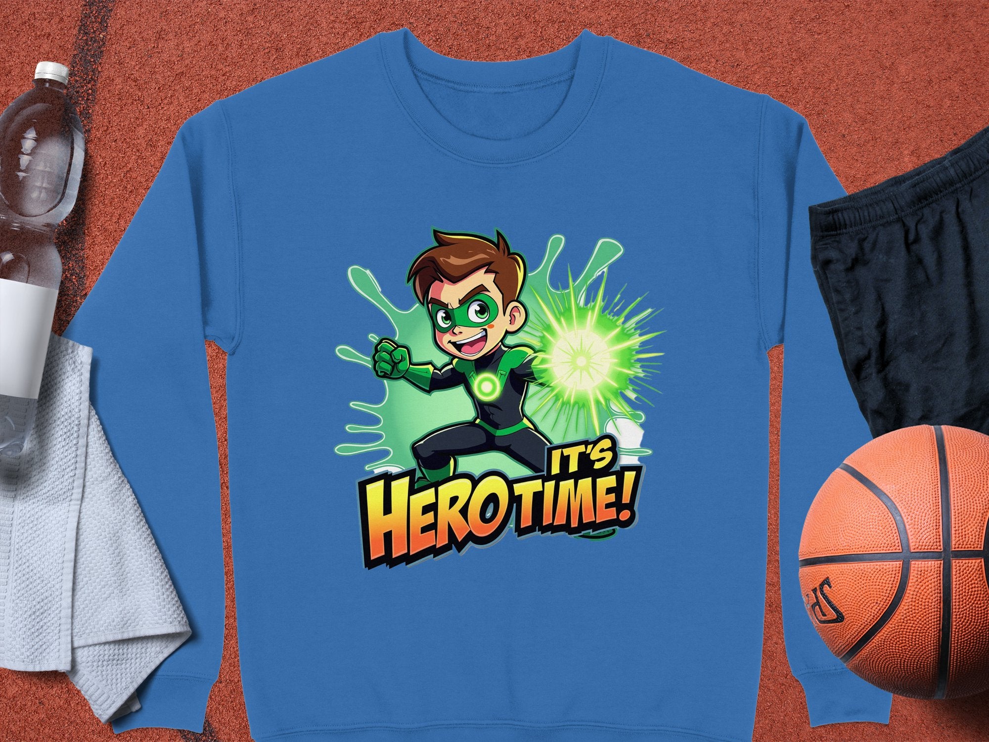 A Garment Graphics Its Herotime sweatshirt, showcasing a cartoon character with a green mask and glowing power, is laid on a surface. Ideal for adventure enthusiasts, its paired with essentials like a water bottle, white towel, black shorts, and an orange basketball.