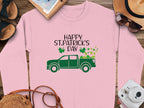 The pink festive sweatshirt by Garment Graphics sports a green truck with shamrocks and clovers, Happy St. Patricks Day above. It is set against a wooden backdrop with shoes, a camera, sunglasses, a straw hat, and a notepad.