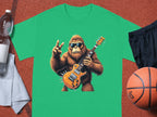 A green T-shirt from Garment Graphics features a gorilla in sunglasses playing an electric guitar. Its displayed among a water bottle, towel, shorts, and basketball on an orange textured surface—ideal for music lovers with a sporty vibe.