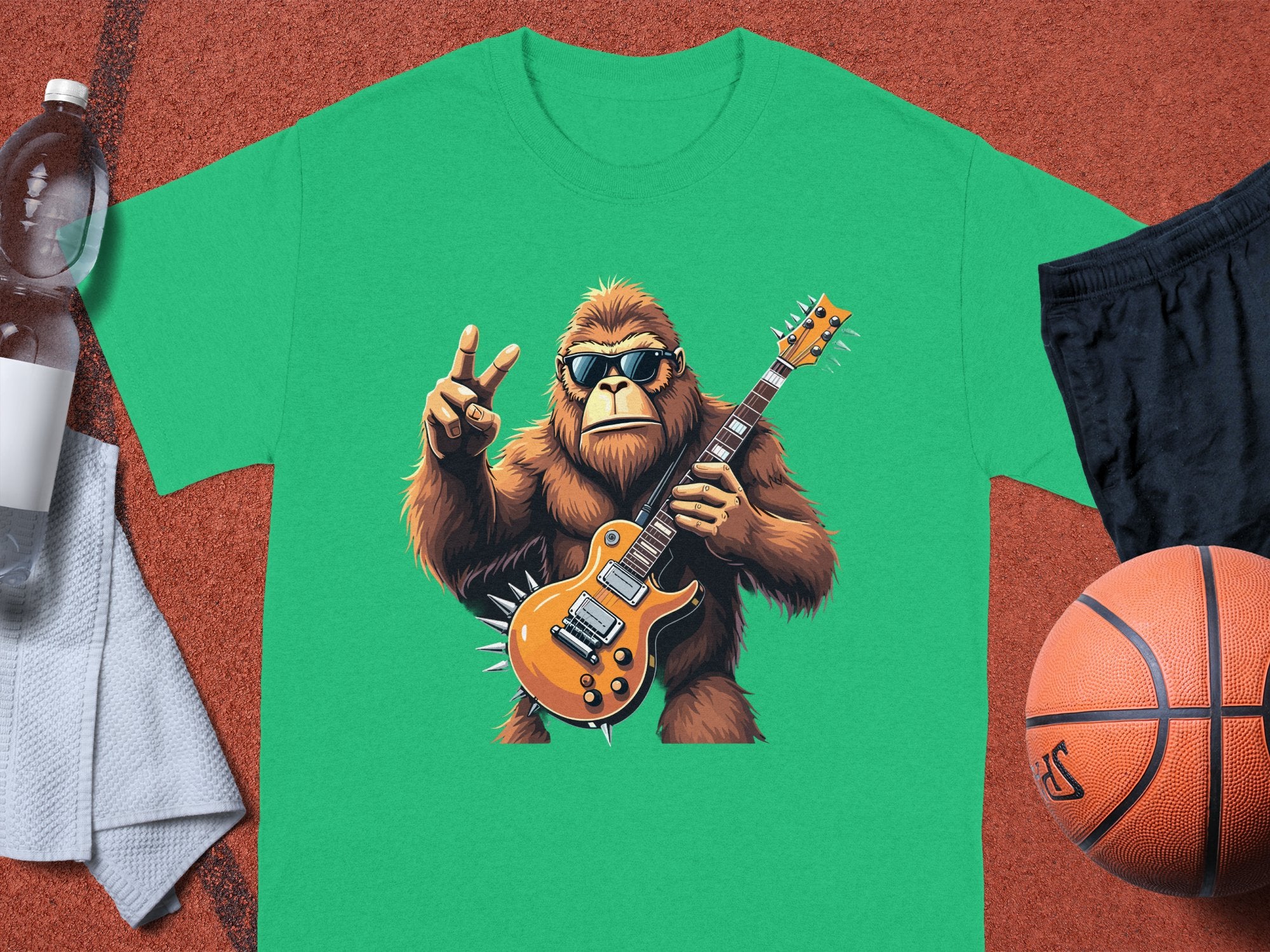 A green T-shirt from Garment Graphics features a gorilla in sunglasses playing an electric guitar. Its displayed among a water bottle, towel, shorts, and basketball on an orange textured surface—ideal for music lovers with a sporty vibe.