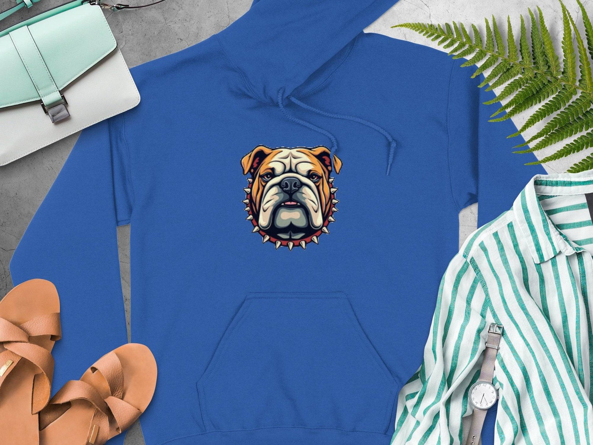 Garment Graphics blue bulldog design hoodie features a cartoon graphic and spiked collar at the center. Pair it with tan sandals, a white and green striped shirt, a watch, a white handbag, and green fern leaves on a gray background for an eye-catching ensemble.