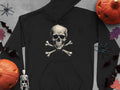 A fashion-forward black hoodie by Garment Graphics, showcasing a skull and crossbones design, is surrounded by Halloween décor—pumpkins, a mini skeleton, bat cutouts, and autumn leaves—artfully arranged on a gray surface.