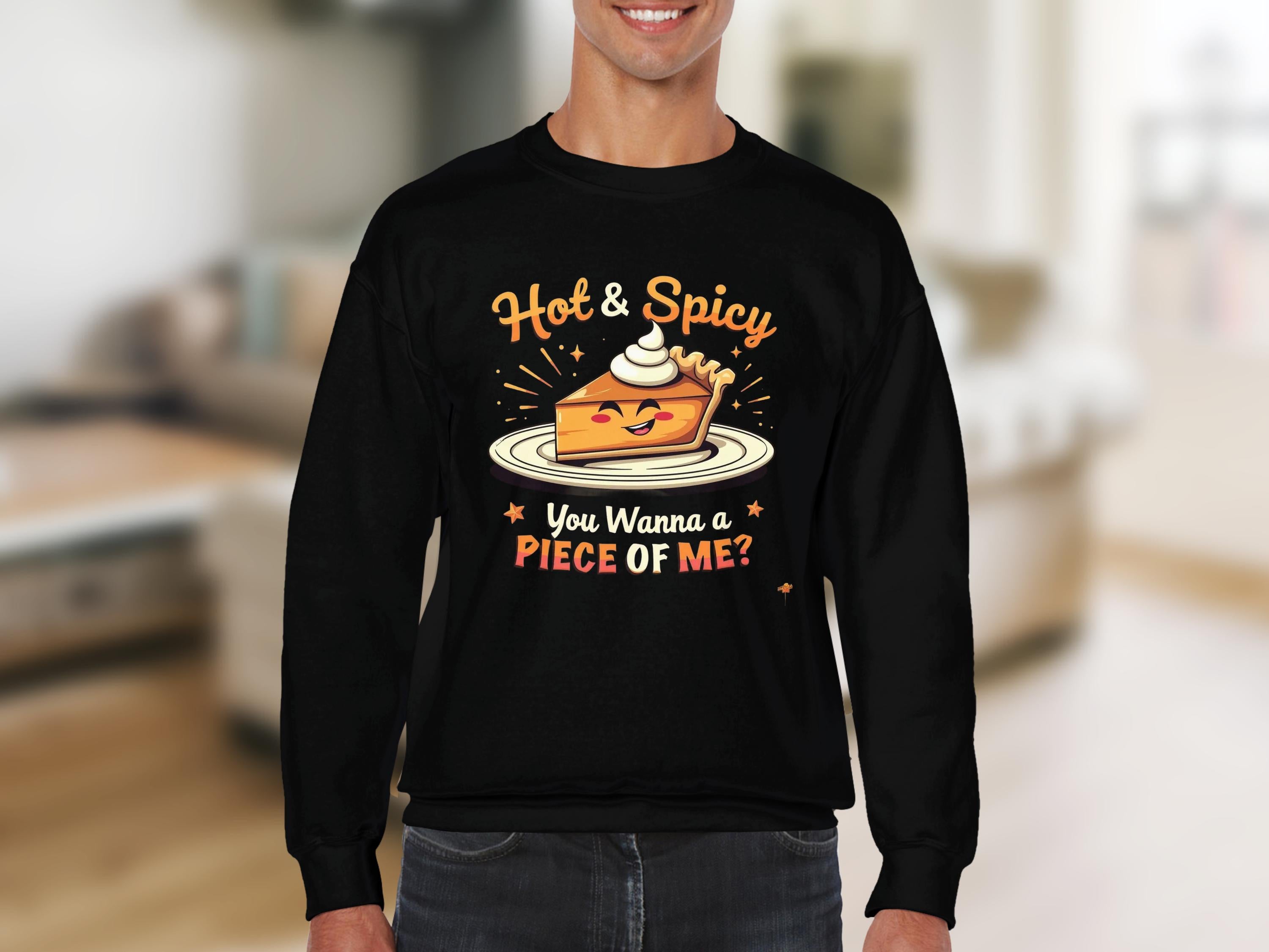 A person wears a black sweatshirt from Garment Graphics Hot and Spicy Sweatshirts collection, featuring a playful pie topped with whipped cream, Hot & Spicy above it, and You Wanna a Piece of Me? below. Its unisex for all to enjoy its charm against a blurred room backdrop.