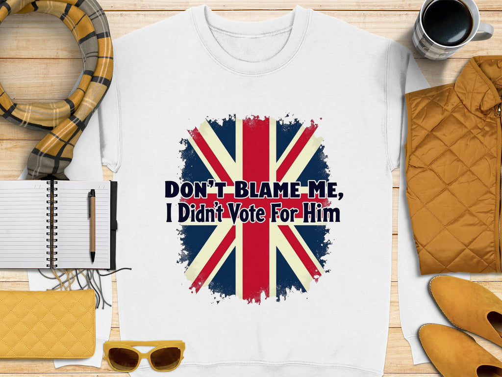 A white unisex t-shirt by Garment Graphics, with a stylized Union Jack and the slogan Dont Blame Me, I Didnt Vote For Him, is displayed alongside a plaid scarf, notebook, pen, yellow clutch, sunglasses, coffee cup, and yellow jacket on a wooden surface.