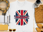 A white unisex t-shirt by Garment Graphics, with a stylized Union Jack and the slogan Dont Blame Me, I Didnt Vote For Him, is displayed alongside a plaid scarf, notebook, pen, yellow clutch, sunglasses, coffee cup, and yellow jacket on a wooden surface.