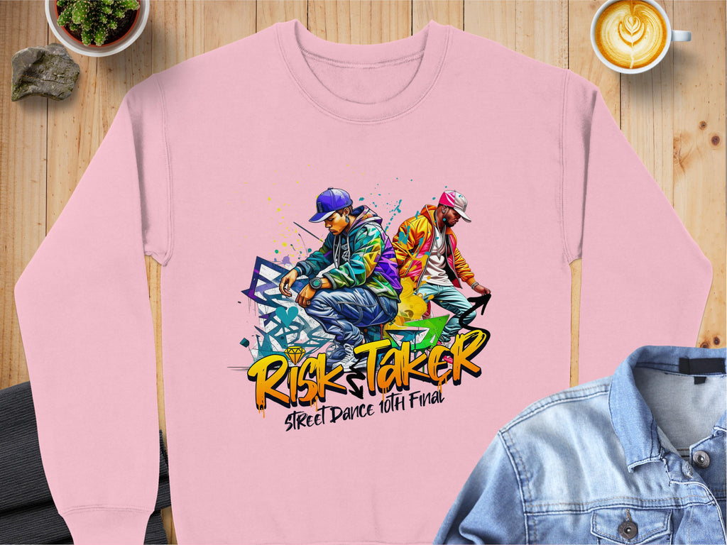 Garment Graphics pink sweatshirt showcases vivid street dance graphics with two dancers in urban outfits against a graffiti backdrop, featuring the text Risk Taker Street Dance 10th Final. Its displayed on a wooden surface alongside a denim jacket and latte.