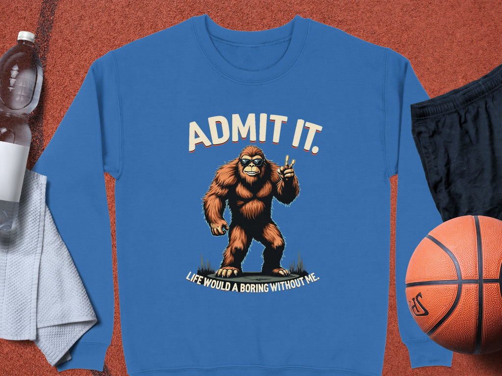 The Garment Graphics Admit It Sweatshirt showcases a blue design with Bigfoot holding a peace sign and the text, Admit it. Life would be boring without me, styled alongside a basketball, black shorts, towel, and water bottle.