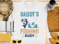 Garment Graphics offers a white Daddys Fishing Buddy t-shirt, highlighting a boat fishing scene. Pair it with a scarf, coffee cup, jacket, notepad and pens, yellow purse, and sunglasses for the perfect fishing enthusiast ensemble on any wooden surface.