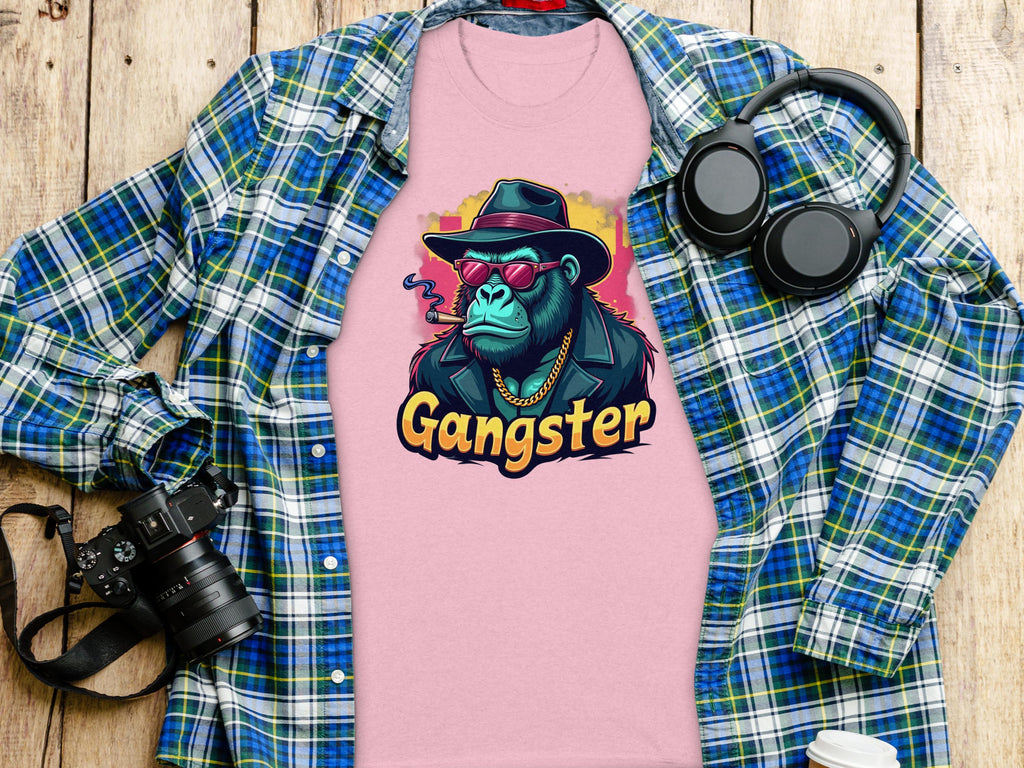 A pink Garment Graphics Gangster Gorilla T-Shirt featuring a gorilla in a hat, sunglasses, and jacket is displayed on a wooden surface alongside a classic-fit blue plaid shirt, headphones, camera, and disposable coffee cup.
