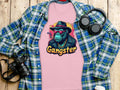 A pink Garment Graphics Gangster Gorilla T-Shirt featuring a gorilla in a hat, sunglasses, and jacket is displayed on a wooden surface alongside a classic-fit blue plaid shirt, headphones, camera, and disposable coffee cup.