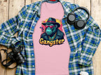 A pink Garment Graphics Gangster Gorilla T-Shirt featuring a gorilla in a hat, sunglasses, and jacket is displayed on a wooden surface alongside a classic-fit blue plaid shirt, headphones, camera, and disposable coffee cup.
